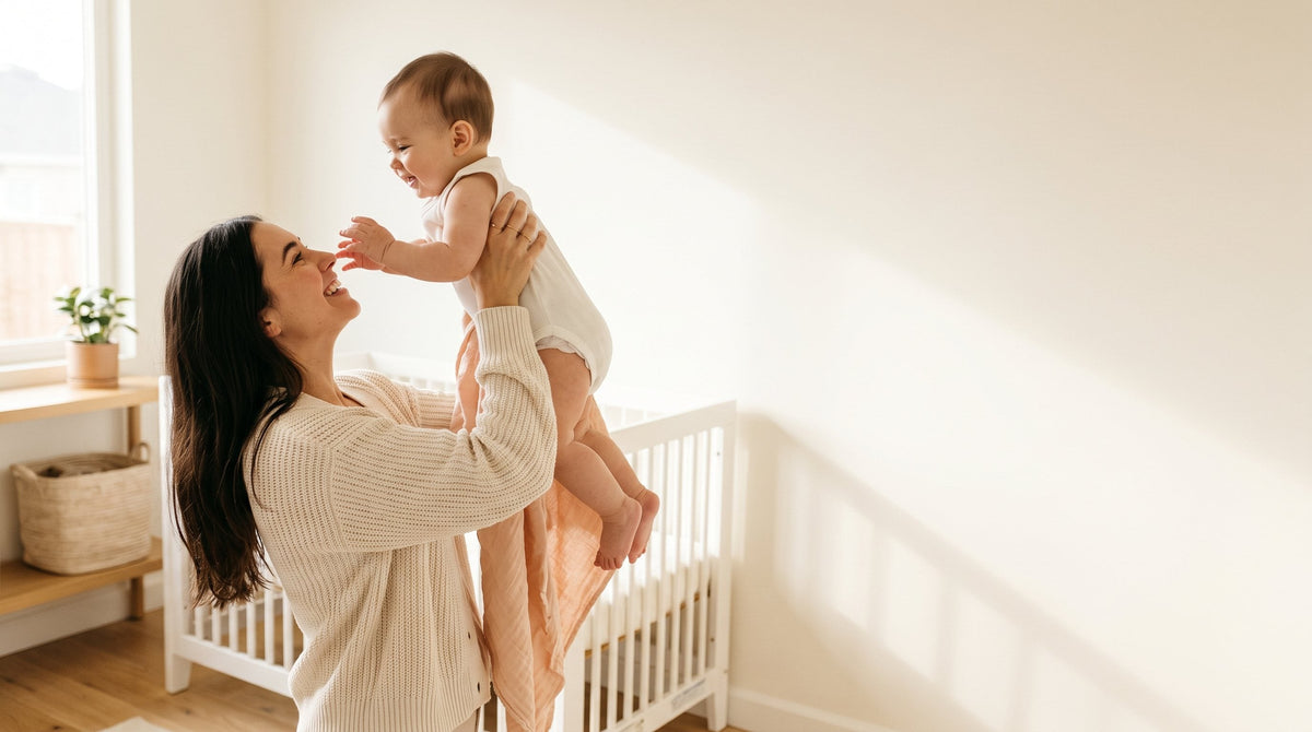 Mother lifting her laughing baby beside a white crib in a sunny nursery