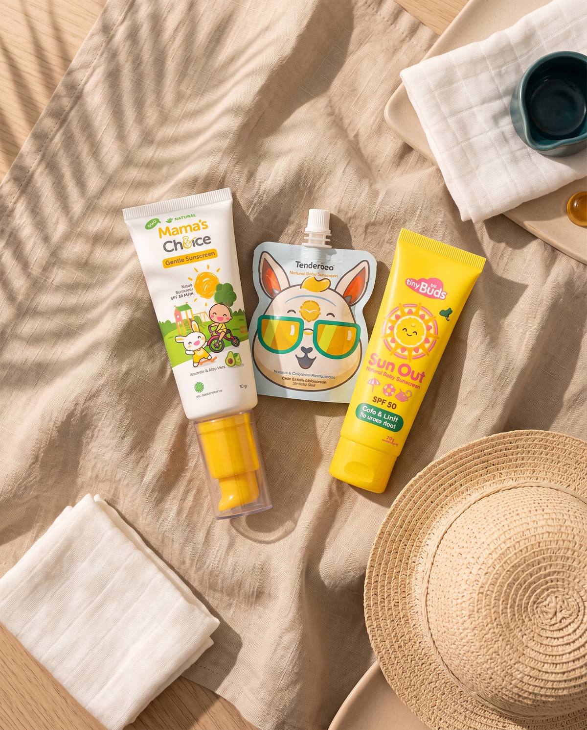 Baby sunscreens on linen with a palm-leaf shadow and a straw sun hat